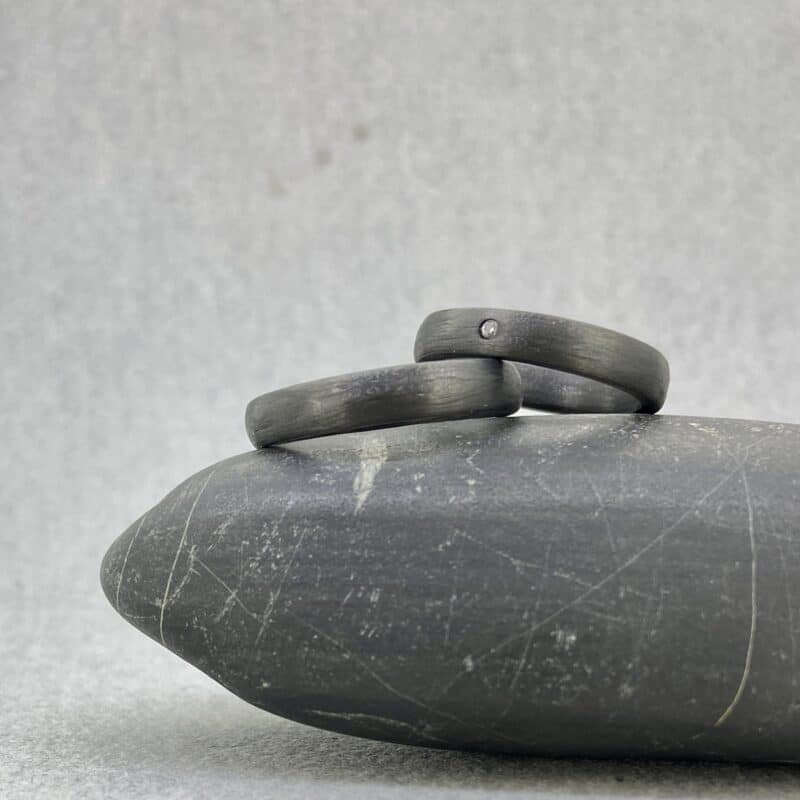 Toma de catálogo en pareja de las alianzas de media caña de carbono negro de 4 mm de Joyería Zayro. Muestra un cambio de perspectiva tridimensional donde una de las alianzas aparece apoyada encima de la otra, resaltando la curvatura exterior del perfil y la textura orgánica mate y opaca del composite.