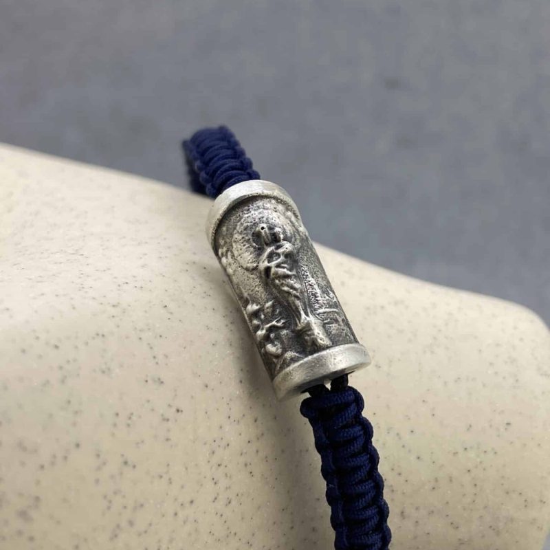 Pulsera de la Virgen del Pilar de Joyería Zayro tejida en hilo náutico de nailon azul con macramé y pieza central de plata de ley, apoyada sobre un jarrón beige de estudio.