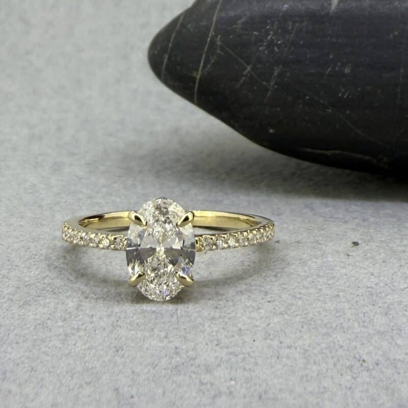 Anillo de compromiso de oro amarillo de 18 quilates con diamante central ovalado de laboratorio y hombros engastados con diamantes brillantes sobre fondo neutro.