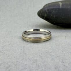 Fotografía original de Joyería Zayro de una alianza individual bicolor de 18 kilates colocada en perspectiva oblicua. El anillo, apoyado sobre una piedra de canto rodado gris oscuro de taller, presenta una banda base de 4 mm de ancho en oro blanco con textura de arena mate satinada y un aro fino central superpuesto de 1 mm en oro amarillo pulido brillo.