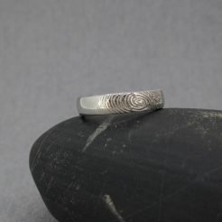 Fotografía original de Joyería Zayro de una alianza individual de oro blanco de 18 kilates colocada de frente en posición vertical. El anillo, de 4 mm de ancho, muestra un perfil geométrico almenado pulido brillo y un grabado láser de huella dactilar en relieve exterior