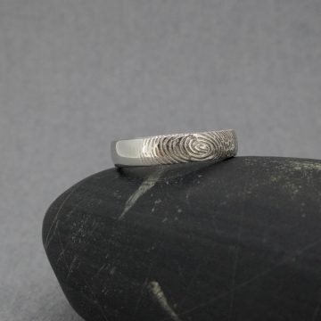 Fotografía original de Joyería Zayro de una alianza individual de oro blanco de 18 kilates colocada de frente en posición vertical. El anillo, de 4 mm de ancho, muestra un perfil geométrico almenado pulido brillo y un grabado láser de huella dactilar en relieve exterior