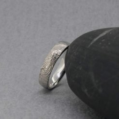 Vista de canto en perspectiva oblicua de una alianza individual de oro blanco de 18kt de Joyería Zayro apoyada en una base de mármol veteado junto a una hoja de eucalipto. La toma resalta el grosor del perfil plano almenado mate, la textura del grabado exterior y el interior del anillo pulido brillo con acabado ergonómico confort.