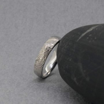 Vista de canto en perspectiva oblicua de una alianza individual de oro blanco de 18kt de Joyería Zayro apoyada en una base de mármol veteado junto a una hoja de eucalipto. La toma resalta el grosor del perfil plano almenado mate, la textura del grabado exterior y el interior del anillo pulido brillo con acabado ergonómico confort.