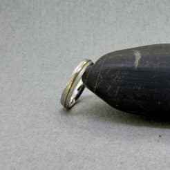 Vista oblicua y de canto de una alianza de oro de 18 quilates de Joyería Zayro apoyada de lado sobre una piedra gris oscura. La toma resalta con nitidez el grosor del perfil plano de oro blanco de 4 mm, el relieve del anillo central de oro amarillo de 1 mm y la cara interna de la banda con un pulido brillo espejo ergonómico confort.