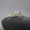 Fotografía original de estudio de Joyería Zayro de una pareja de alianzas de boda de oro blanco de 18 kilates con perfil plano y 4 mm de ancho. Los dos anillos están dispuestos juntos sobre un canto rodado gris ante un fondo claro, mostrando un acabado texturizado tipo corteza de árbol; la alianza de caballero es lisa y la de señora lleva cinco diamantes blancos brillantes engastados en hilera en el centro superior de la banda recta.