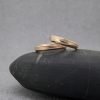 Fotografía original de Joyería Zayro que muestra una pareja de alianzas de boda de oro rosa de media caña. Los anillos están dispuestos juntos sobre un canto rodado gris oscuro ante un fondo gris claro, destacando el diseño exclusivo de Jorge de Roque con una mitad de textura arena mate y otra mitad pulida brillo, divididas por una línea diagonal.
