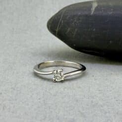 Fotografía de bodegón artesanal en el taller de Joyería Zayro; el anillo solitario asimétrico Hermes en oro blanco brilla sobre una superficie natural de canto rodado gris, destacando el corte del diamante natural y las líneas limpias de la orfebrería de autor.