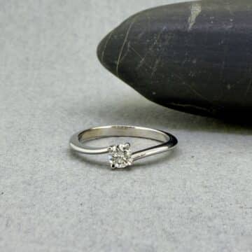 Fotografía de bodegón artesanal en el taller de Joyería Zayro; el anillo solitario asimétrico Hermes en oro blanco brilla sobre una superficie natural de canto rodado gris, destacando el corte del diamante natural y las líneas limpias de la orfebrería de autor.