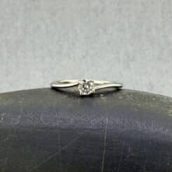 Anillo de compromiso solitario Hermes en oro blanco de 18k con un diamante natural central de 0.20 cts certificado por GIA; diseño asimétrico con brazos desalineados pulidos, expuesto sobre una piedra de canto rodado gris en el taller de Joyería Zayro en Zaragoza.