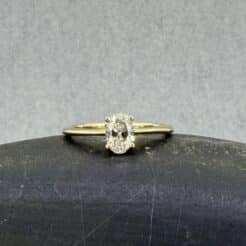 Anillo de compromiso solitario en oro amarillo de 18k con un diamante de laboratorio central de talla oval con montura Venus de cuatro garras y brazo en cuchilla, sobre fondo blanco de catálogo.