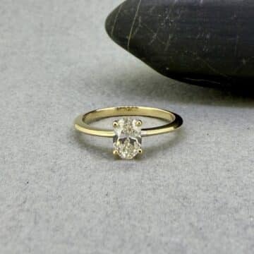 Anillo de compromiso solitario Venus en oro amarillo con un diamante de laboratorio oval central, expuesto sobre la astillera de madera texturizada del taller de Joyería Zayro en Zaragoza, mostrando la escala real de la joya junto a sutiles marcas de herramientas artesanales.